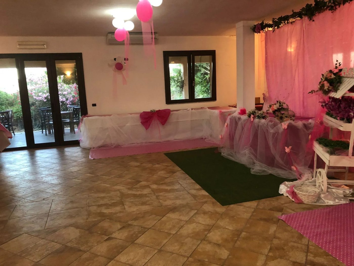 Sala con tavoli drappeggiati in tulle bianco e rosa, palloncini fucsia, fiori e vetrata sul giardino dell'Agriturismo Monte Alma